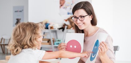 teacher holding a smiley and frowny face and child pointing at the smile